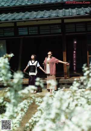Two young women standing next to each other in front of bushes.